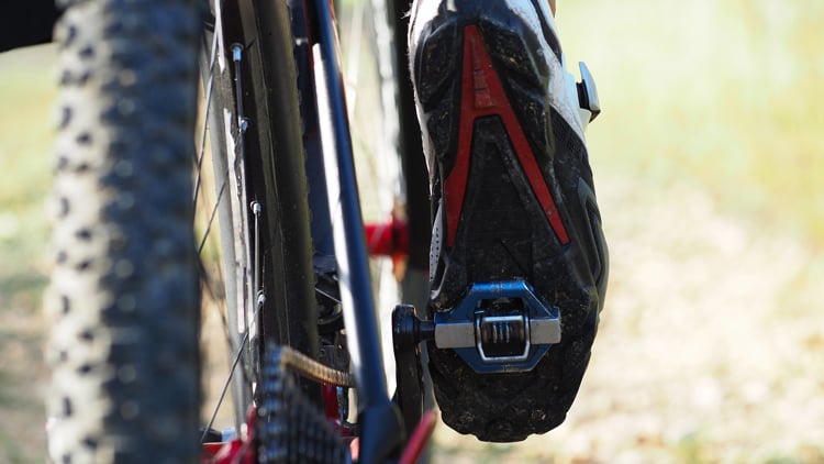 Nahaufnahme eines Fahrradpedals mit eingeklicktem Radschuh; Fokus auf die Verbindung zwischen Schuh und Pedal, unscharfer Hintergrund deutet auf eine Outdoor-Umgebung hin.