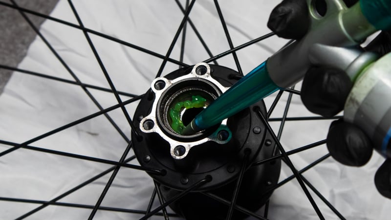 Nahaufnahme der Hand eines Fahrradmechanikers mit schwarzem Latexhandschuh beim Fetten der Hinterradnabe mit einer Fettpresse.