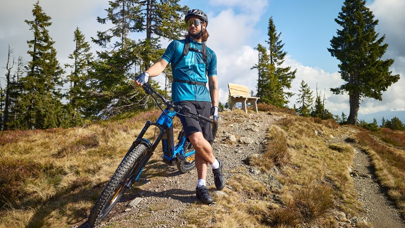 Ein Mann in Fahrradkleidung steht mit einem Mountainbike auf einem Bergpfad. Im Hintergrund befinden sich Bäume, ein Holzbänkchen und ein teilweise bewölkter blauer Himmel.