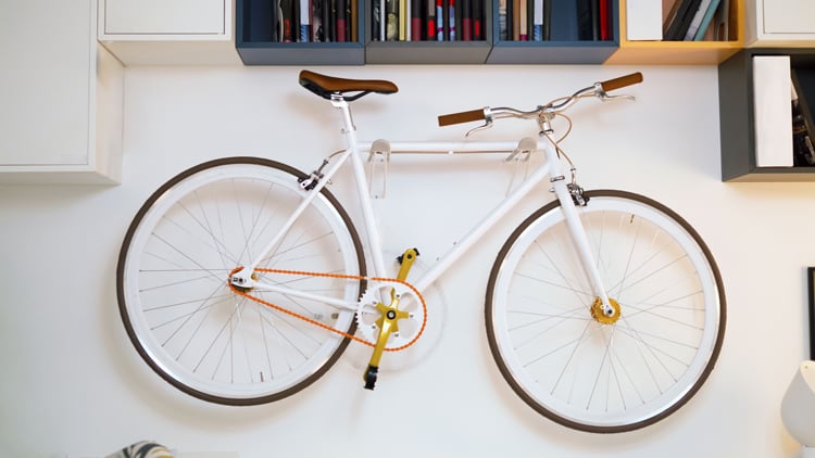 Weißes Fahrrad mit braunem Lenker und braunem Sattel, orangefarbener Kette und gelben Pedalen, an einer Wand montiert; darüber Regale mit Büchern und Gegenständen.