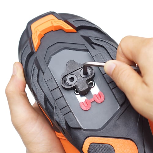 Nahaufnahme einer schwarzen und orangefarbenen Radschuhsohle, an der mit einem Inbusschlüssel eine schwarze Cleat mit roten Komponenten befestigt oder justiert wird.