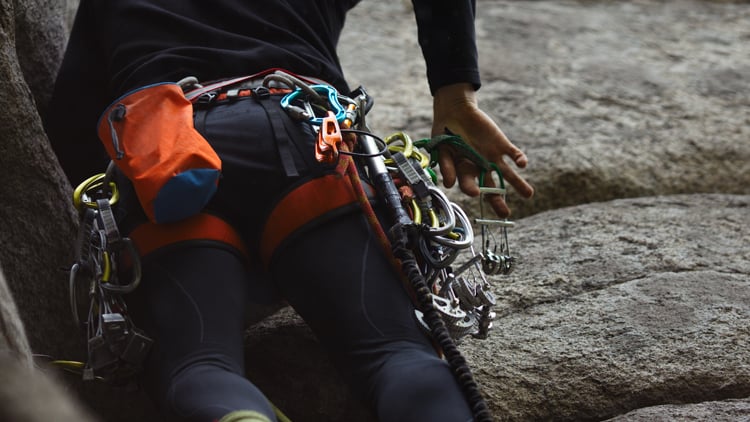 Eine Person beim Felsklettern, ausgestattet mit Klettergurt, Karabinern, Express-Sets und weiterem Kletterequipment. Eine Hand greift nach der Felsoberfläche.