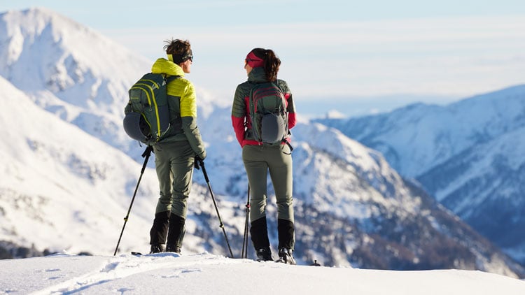 Zwei Personen stehen auf einer verschneiten Berglandschaft, ausgerüstet mit Rucksäcken und Wanderstöcken. Sie tragen Winterkleidung mit Jacken, Hosen und Stiefeln. Im Hintergrund sind schneebedeckte Berge unter klarem Himmel zu sehen