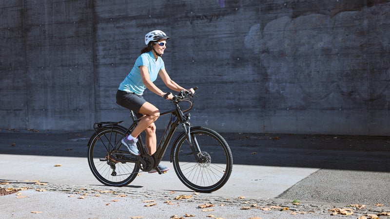 Person auf einem e-Bike auf einem asphaltierten Weg. Sie trägt einen Helm, ein hellblaues Shirt, schwarze Shorts und Sportschuhe. Im Hintergrund eine Betonwand mit Graffiti und herabgefallenes Laub auf dem Boden.
