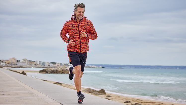 Mann joggt auf einer Promenade am Meer in roter Tarnjacke und schwarzen Shorts.