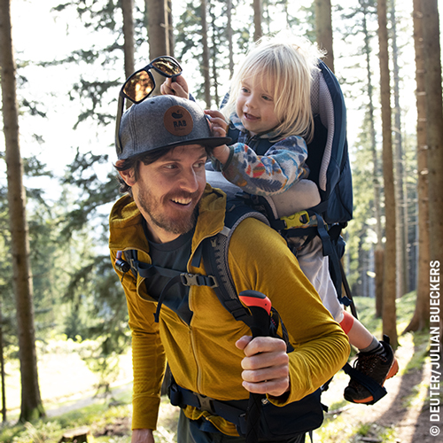 Erwachsene Person mit Kind in Kindertragerucksack wandert durch den Wald; das Kind greift nach dem Hut der Person.