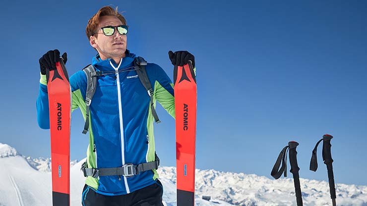 Get Green Person steht auf einem verschneiten Berg und hält zwei rot-schwarze Skier der Marke ‚ATOMIC‘. Sie trägt eine blau-grüne Jacke, schwarze Handschuhe und einen Rucksack. Rechts im Schnee stecken zwei Skistöcke. Im Hintergrund sind schneebedeckte Berge unter klarem blauem Himmel zu sehen