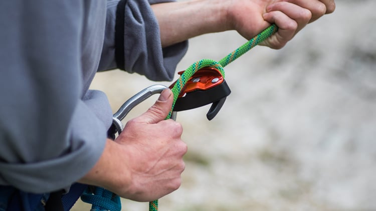 Eine Person mit grauem Langarmshirt hält ein grünes Kletterseil mit beiden Händen. Das Seil ist durch ein orange-schwarzes Sicherungsgerät geführt, das die Person fest umgreift. Im Hintergrund ist eine Outdoor-Umgebung zu erkennen, vermutlich ein Klettergebiet.