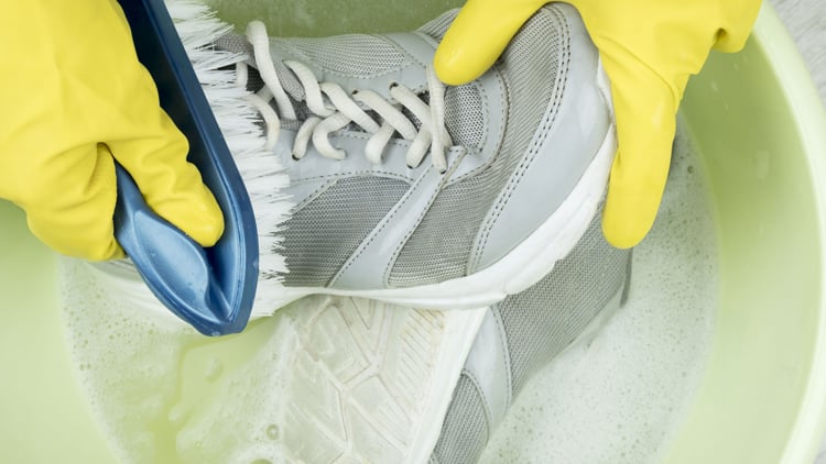 Laufberater Ein Paar Hände mit gelben Gummihandschuhen reinigt einen grau-weißen Turnschuh mit einer blauen Bürste in einem hellgrünen Becken mit Seifenwasser