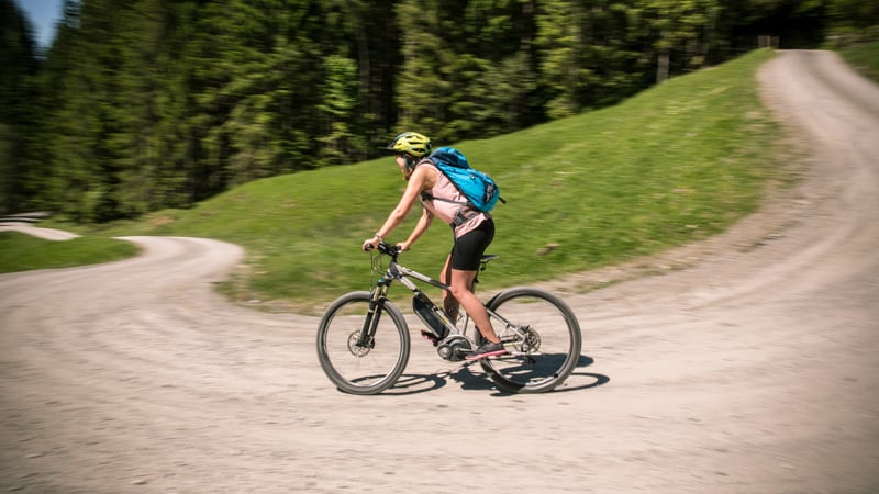 Person mit Helm, Shorts, ärmellosem Oberteil und blauem Rucksack fährt mit dem Fahrrad auf einem Waldweg bergab.