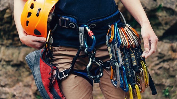 Eine Person mit Kletterausrüstung hält einen orangefarbenen Kletterhelm in der Hand. Am Gurt ist ein roter Kletterschuh befestigt, ebenso wie mehrere Karabiner und Express-Sets. Im Hintergrund ist eine felsige Outdoor-Umgebung zu sehen