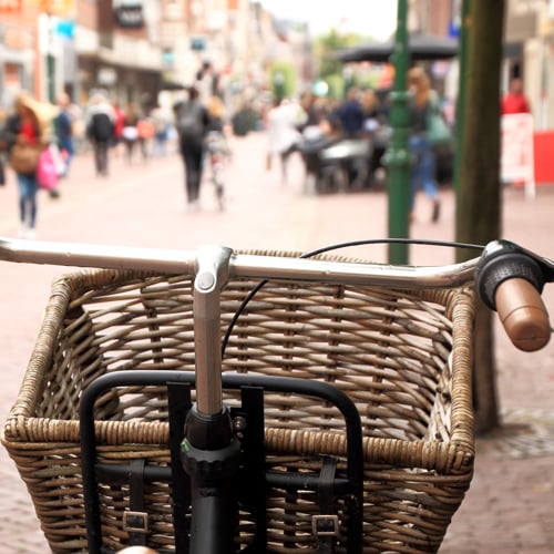 Frontansicht eines Fahrrads mit Weidenkorb am Lenker; im Hintergrund eine belebte Einkaufsstraße mit Passanten und Gebäuden, leicht unscharf dargestellt.