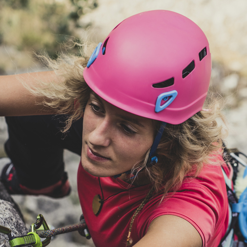 Eine Person beim Outdoor-Klettern mit pinkem Helm und rotem Shirt. Der Hintergrund zeigt eine felsige Oberfläche mit etwas Grünbewuchs.