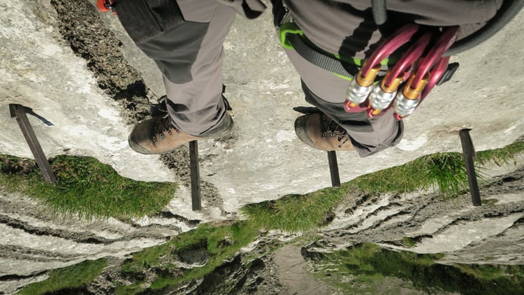 Blick aus der Perspektive eines Kletterers auf einer Klettersteig. Die Füße stehen auf Metalltritten in einer senkrechten Felswand.