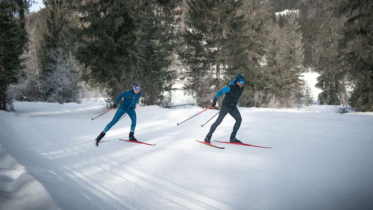 Zwei Personen beim Langlaufen auf einem verschneiten Weg, umgeben von Bäumen. Beide tragen Winter-Sportkleidung mit Jacken, Hosen, Handschuhen und Mützen. Die linke Person ist in Blau gekleidet, die rechte in Schwarz mit blauen Akzenten