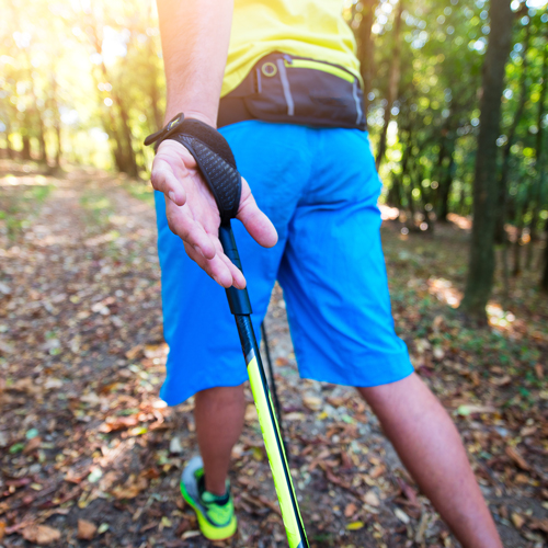 Eine Person beim Nordic Walking auf einem mit Laub bedeckten Waldweg. Sie trägt blaue Shorts, ein gelbes Shirt und grüne Laufschuhe. Sonnenlicht fällt durch die Bäume im Hintergrund