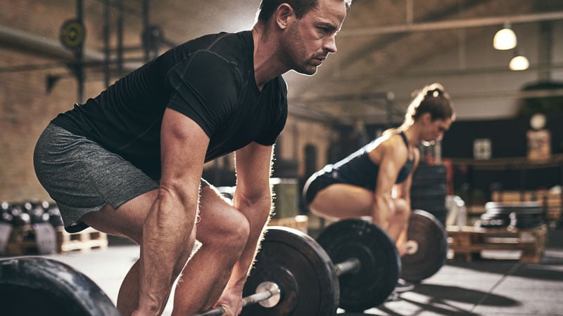 Zwei Personen in einem Fitnessstudio bereiten sich darauf vor, Langhanteln vom Boden zu heben. Die vordere Person trägt ein schwarzes Shirt und graue Shorts, die hintere eine dunkle Trainingskleidung. Im Raum sind Gewichte und Trainingsgeräte verteilt