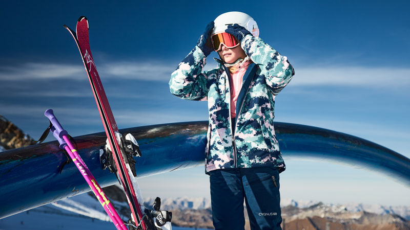 Kind in Winterkleidung mit Helm und Handschuhen steht im Freien vor einer verschneiten Berglandschaft. Das Gesicht ist nicht sichtbar. Neben der Person lehnen zwei Paar Skier und Skistöcke an einem Geländer