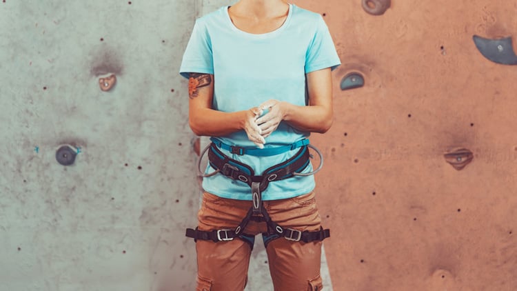 Person in blauem T-Shirt und brauner Hose steht vor einer Kletterwand in einer Halle. Sie trägt einen Klettergurt und bestäubt ihre Hände mit Chalk. Die Wand im Hintergrund ist grau und orange und mit verschiedenen Klettergriffen ausgestattet