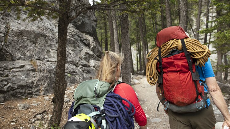 Zwei Personen gehen auf einem Waldpfad mit Kletterausrüstung. Die linke Person trägt ein rotes Shirt und einen grünen Rucksack mit gelbem Helm und blauer Seilschlaufe.