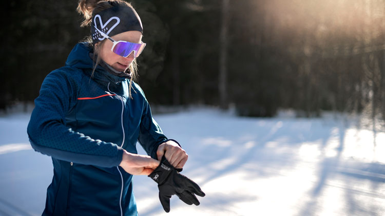 Eine Person steht in einer verschneiten Umgebung im Freien, trägt eine blaue Jacke und richtet schwarze Handschuhe. Das Gesicht ist durch ein violettes Quadrat verdeckt. Im Hintergrund sind Bäume und Sonnenlicht zu sehen, das Schatten auf den Schnee wirft