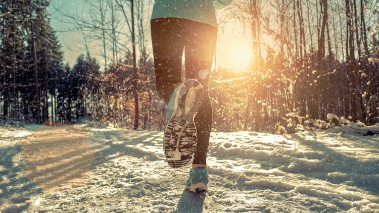 Frau von hinten joggt in sportlicher Kleidung auf einem verschneiten Waldweg bei Sonnenschein.