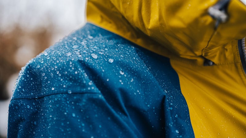 Skibekleidung imprägnieren Nahaufnahme einer Person mit einer wasserabweisenden Jacke in Blau und Gelb, auf deren Oberfläche Wassertropfen zu sehen sind