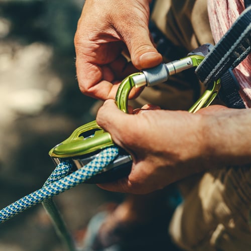 Nahaufnahme von Händen, die einen grünen Karabiner halten und ein blau-weiß gemustertes Kletterseil hindurchführen. Der Hintergrund ist unscharf und lenkt den Fokus auf das Kletterequipment.