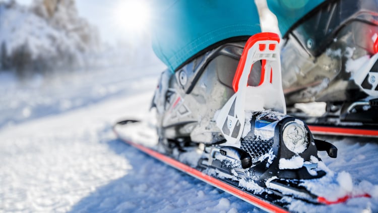 Nahaufnahme von Skischuhen, die in Skibindungen auf Skiern eingerastet sind. Im Hintergrund ist eine verschneite Winterlandschaft mit unscharfen Bäumen und Sonnenlicht zu sehen.