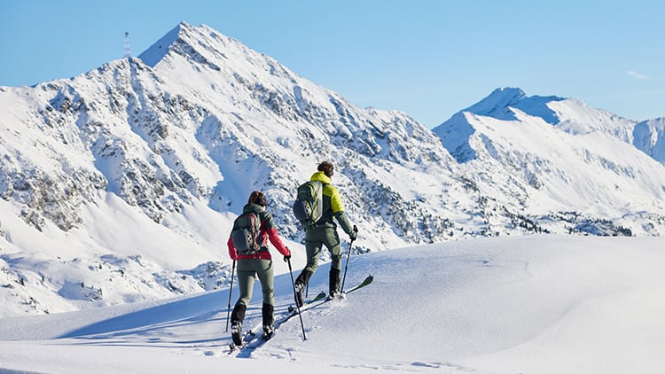Zwei Personen gehen auf einem schneebedeckten Berg eine Skitour. Sie benutzen Skistöcke und tragen Rucksäcke. Im Hintergrund ist eine Reihe von schneebedeckten Bergen unter einem klaren blauen Himmel zu sehen.
