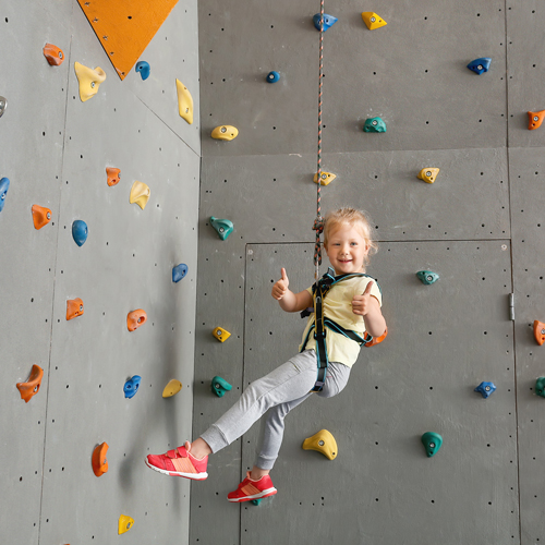 Eine Person klettert an einer Indoor-Kletterwand mit bunten Griffen in Blau, Gelb, Orange und Grün. Sie trägt einen Klettergurt, ist am Seil gesichert und zeigt einen Daumen nach oben.