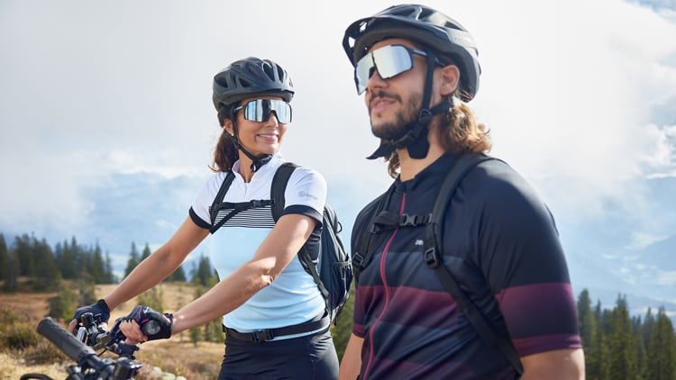 Zwei Personen mit Fahrradhelmen und Radbekleidung stehen mit ihren Fahrrädern in einer landschaftlich reizvollen Umgebung mit Bergen, Bäumen und teils bewölktem Himmel im Hintergrund.