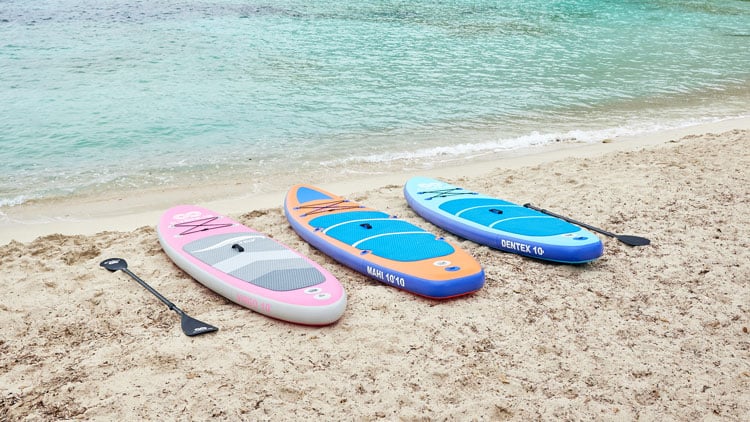 Drei Paddleboards liegen nebeneinander auf einem Sandstrand, mit den Spitzen zum Meer gerichtet. Die Boards sind pink-grau, orange-blau und blau-hellblau gefärbt. Neben jedem Board liegt ein Paddel. Im Hintergrund ist das ruhige Meer mit kleinen Wellen zu sehen