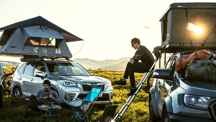 Campingplatz mit zwei Autos auf einer Wiese, beide mit aufgebauten Dachzelten. Eine Person sitzt auf einer Leiter zum Zelt, eine weitere in einem Campingstuhl. Die Szene spielt bei Sonnenauf- oder -untergang mit warmem Licht und Bergen im Hintergrund.