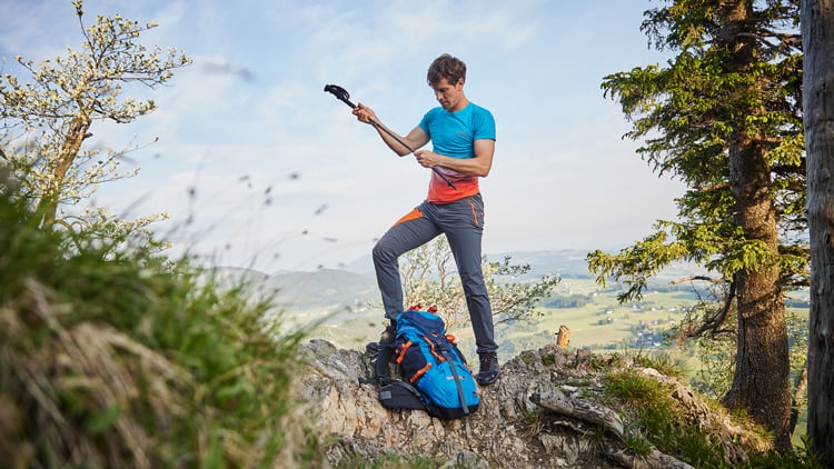 Outdoorberater Wanderer mit Wanderstock auf Felsen, daneben blauer Rucksack, im Hintergrund bewaldete Hügel.