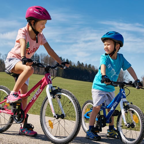 Zwei Kinder fahren auf einem gepflasterten Weg durch ein offenes Feld Fahrrad. Das Kind links trägt einen pinken Helm und fährt ein pinkes Fahrrad, das Kind rechts trägt einen blauen Helm und fährt ein blaues Fahrrad. Im Hintergrund sind Bäume zu sehen