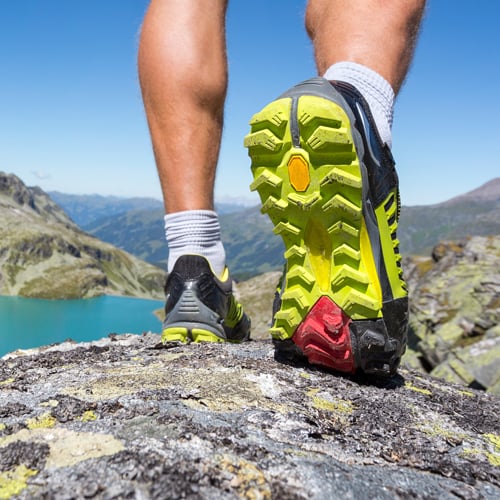 Nahaufnahme von Beinen und Sportschuhen einer Person auf felsigem Untergrund; Fokus auf die profilierte Schuhsohle mit gelben und roten Akzenten, im Hintergrund Berge und ein See unter klarem Himmel