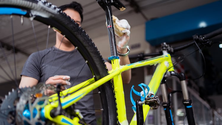 Person reinigt ein gelbes Fahrrad mit blauen Akzenten; wischt mit einem Tuch den Sattelstützbereich, im Hintergrund eine Werkstatt.