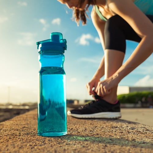 Nahaufnahme einer blauen Wasserflasche auf Beton; im Hintergrund bindet eine Person mit schwarzer Sportkleidung und türkisfarbenem Oberteil die Schnürsenkel ihrer schwarzen Sportschuhe unter freiem Himmel