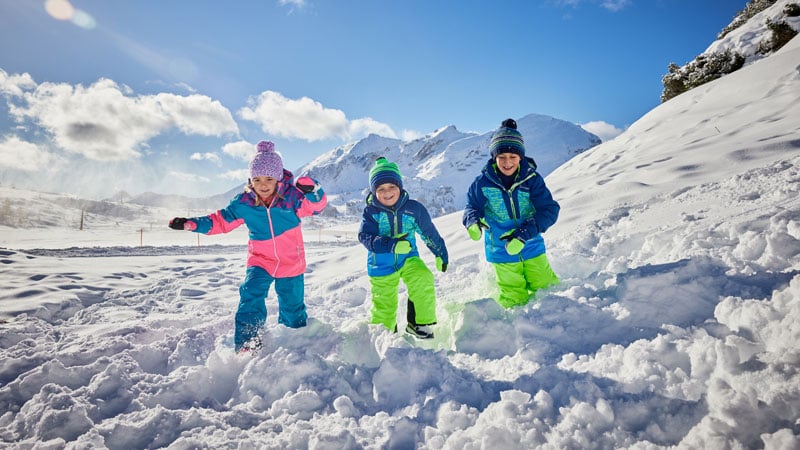 Drei Kinder in bunter Winterkleidung spielen im Schnee vor einer verschneiten Berglandschaft mit klarem, blauem Himmel und vereinzelten Wolken. Das linke Kind trägt ein pink-blaues Outfit, das mittlere ein grün-blaues und das rechte ein blau-grünes