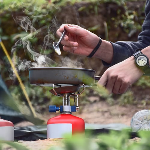 Person kocht im Freien auf einem tragbaren Gaskocher mit roter Kartusche. Eine Hand hält einen Löffel, die andere den Pfannengriff. Aus der Pfanne steigt Dampf auf. Die Person trägt eine Armbanduhr, im Hintergrund sind Pflanzen und Grün zu sehen