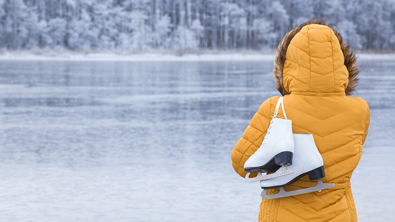 Eine Frau in gelber Winterjacke trägt weiße Schlittschuhe über der Schulter an einem zugefrorenen See.