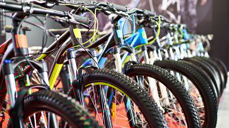 Reihe von Mountainbikes mit dicken, grobstolligen Reifen und farbigen Metallrahmen (u. a. blau, gelb, rot); die Lenker sind mit Bremshebeln und Schalthebeln ausgestattet, im Hintergrund unscharfe Innenraumatmosphäre.