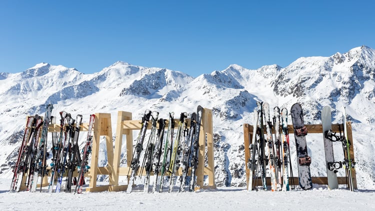 Reihe von Skiern und Snowboards, die auf einem verschneiten Berghang aufrecht in Holzständern stehen. Im Hintergrund sind schneebedeckte Berge unter klarem, blauem Himmel zu sehen