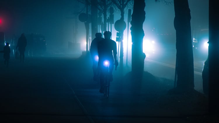 Zwei Radfahrer fahren bei Nacht durch eine neblige Szenerie auf einem beleuchteten Weg; ihre Fahrradlichter leuchten den Weg aus, im Hintergrund sind Bäume, Straßenlaternen mit blauem Lichtschein, Fußgänger und Autoscheinwerfer zu sehen.