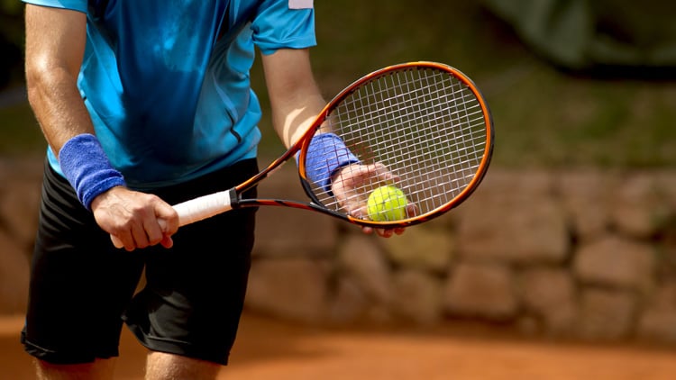 Person in blauem Shirt und schwarzen Shorts hält einen Tennisschläger mit orangem Rahmen und bereitet sich darauf vor, einen gelben Tennisball zu schlagen. Im Hintergrund sind eine Steinmauer und etwas Grün zu sehen