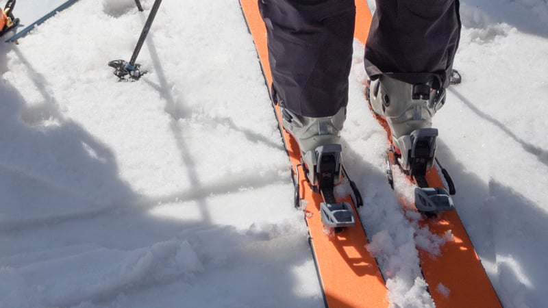 Ein Tourengeher steht mit orangefarbenen Tourenski und Skischuhen im Schnee.