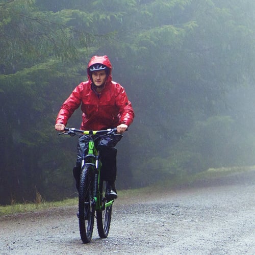 Person mit roter Regenjacke und schwarzer Regenhose fährt im Regen auf einem Waldweg Fahrrad