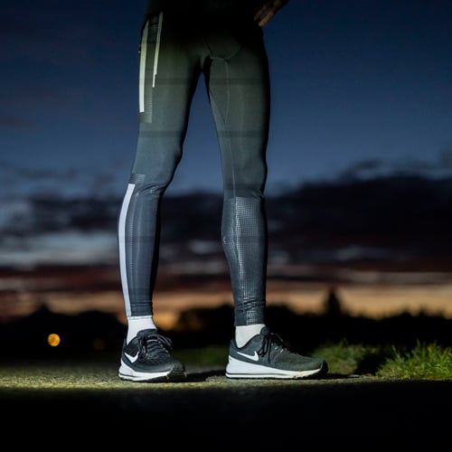 Untere Körperhälfte einer Person in dunklen Sportleggings und schwarzen Laufschuhen mit weißen Sohlen, stehend im Freien bei Sonnenauf- oder -untergang mit wolkigem Himmel und orangefarbenem Horizont