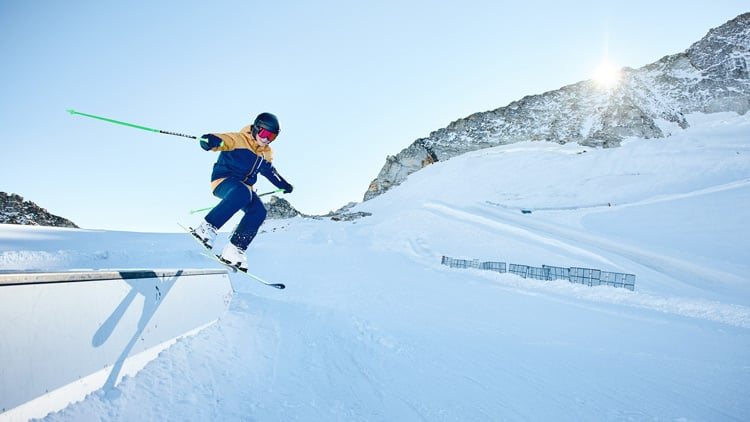Kind in Wintersportausrüstung mit Helm und Skibrille führt einen Sprung oder Trick auf einem verschneiten Berghang aus. Er befindet sich in der Luft und hält Skistöcke. Im Hintergrund sind schneebedeckte Berge zu sehen, über einem Gipfel scheint die Sonne hell
